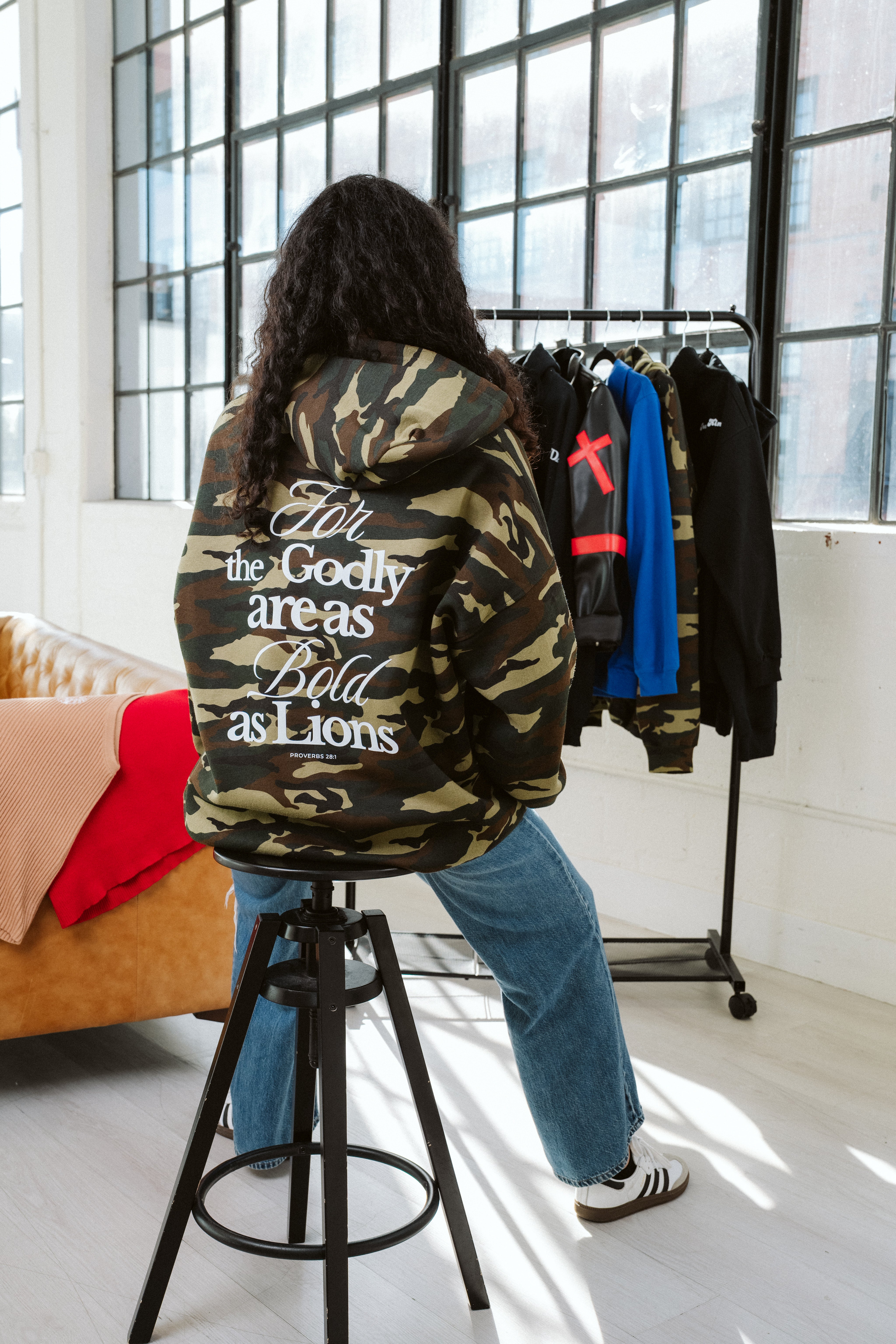 Person wearing a camouflage hoodie with text, standing in a room with clothes on a rack.