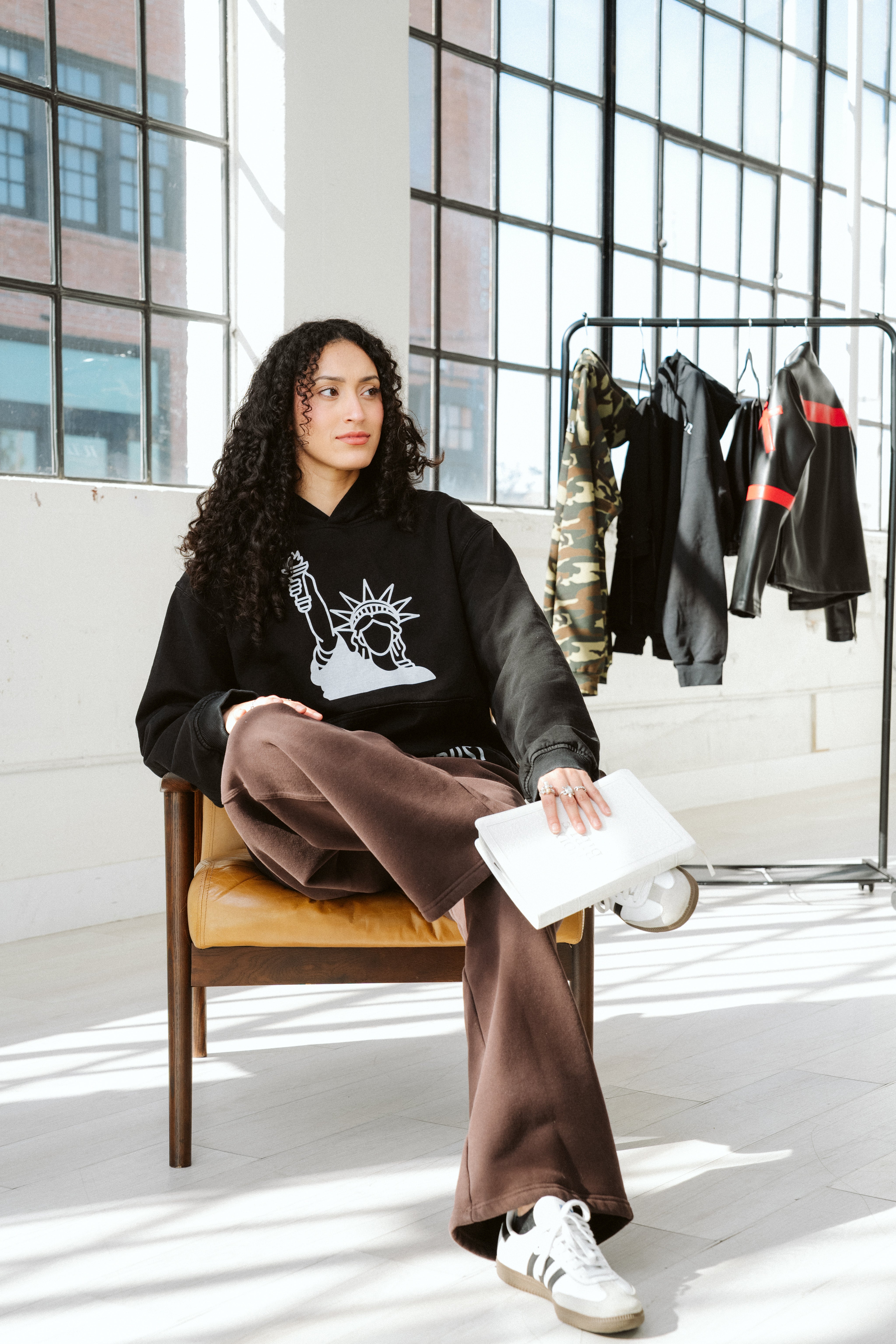 Woman sitting on a chair wearing a black hoodie with a graphic design, brown pants, and white sneakers in a modern indoor setting.