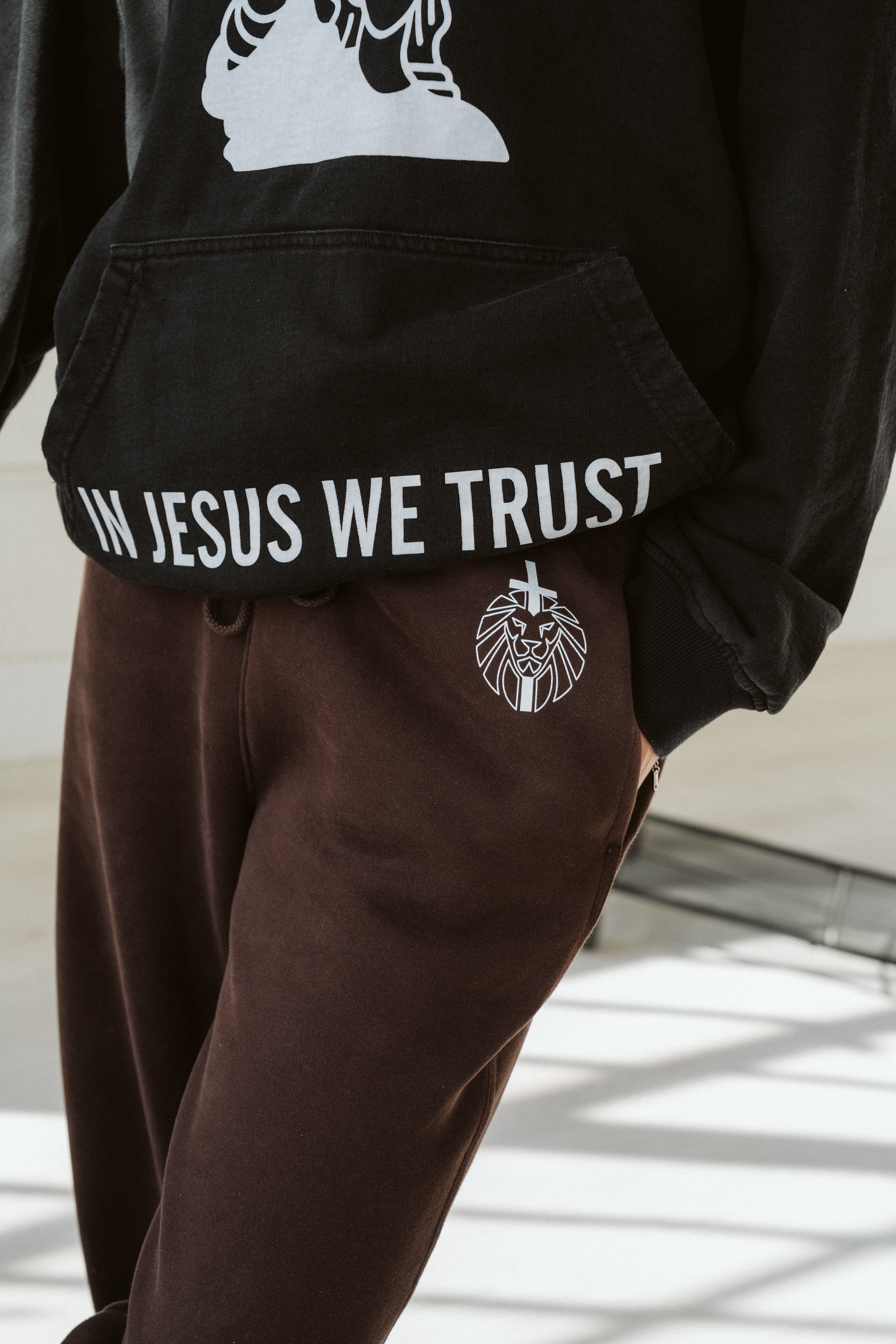 Person wearing a black hoodie with 'IN JESUS WE TRUST' text and brown pants.