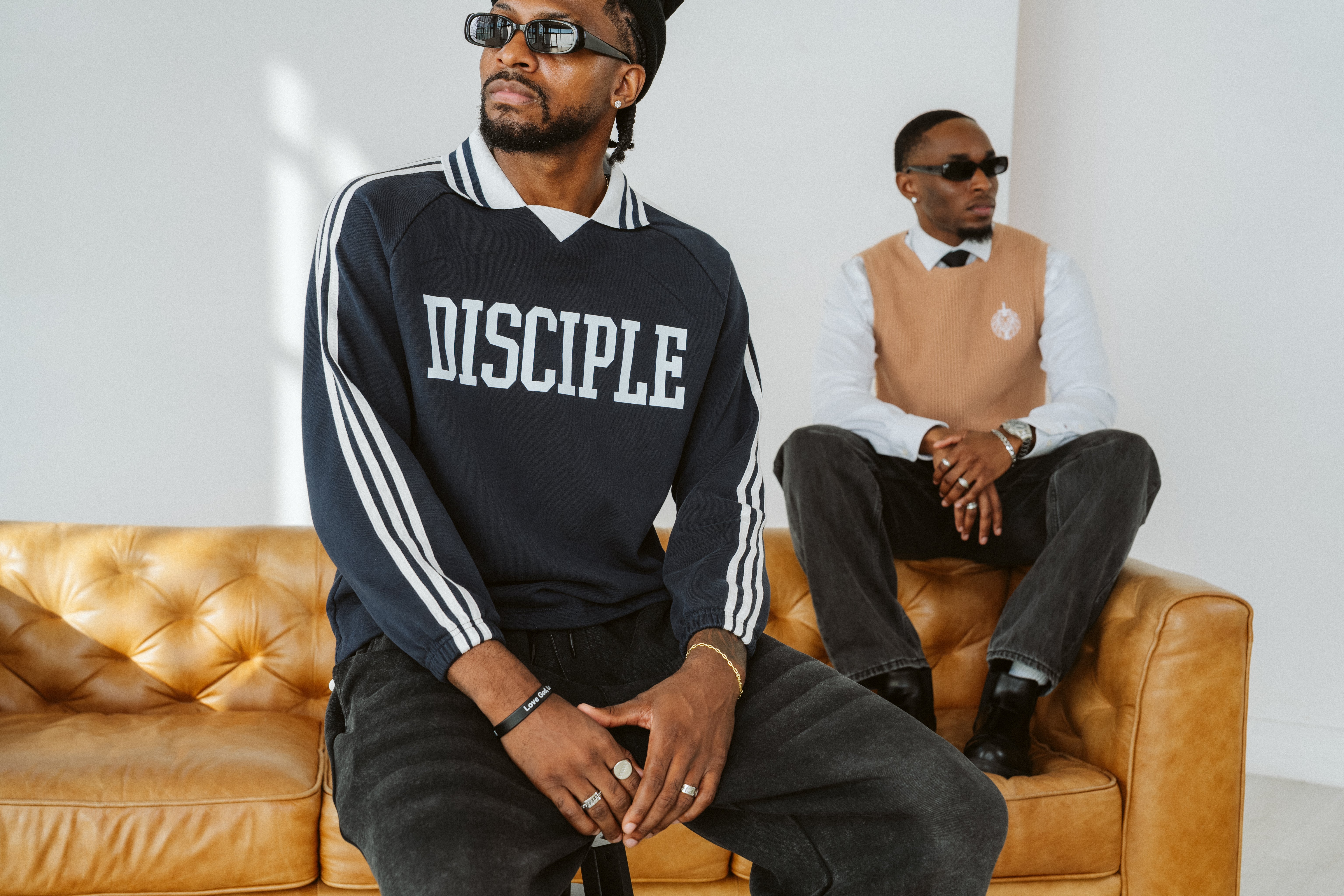 Two men sitting on a brown leather couch, one wearing a 'Disciple' sweatshirt.